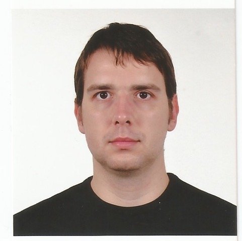 Portrait of a man with short dark hair, wearing a black shirt, facing forward against a plain white background.