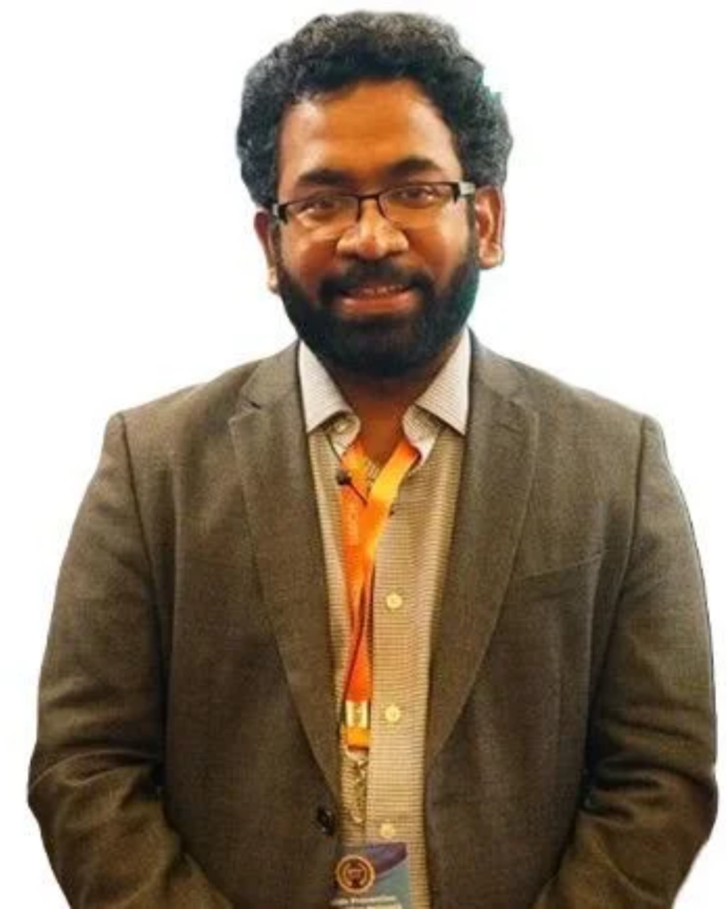 Smiling man with glasses and beard in a brown blazer, orange lanyard visible, standing against a white background.