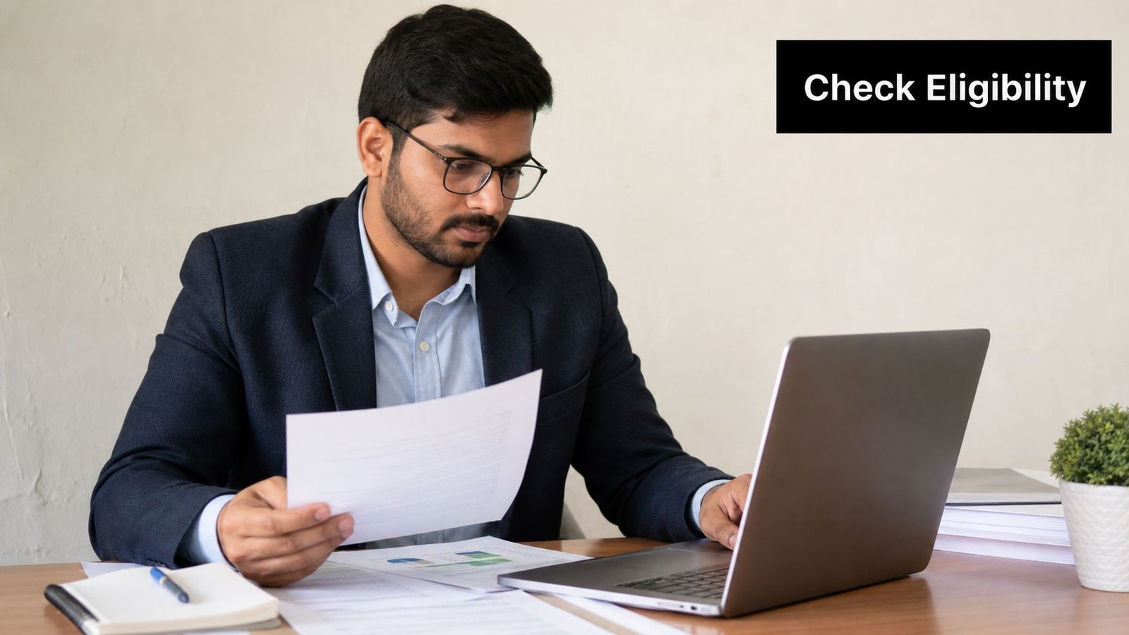 A professional man in a suit reviewing documents while working on a laptop at his desk.