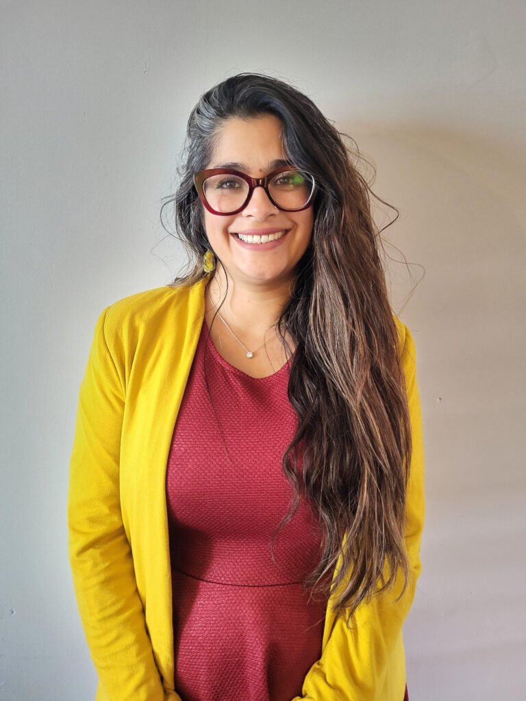 Smiling woman with long dark hair and large red-framed glasses, wearing a yellow cardigan over a red top, against a gray wall.